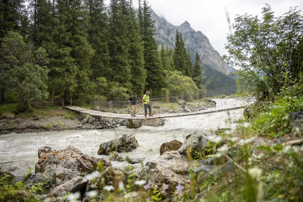 Two hikers crossing a wooden suspension bridge over a river in Kyrgyzstan